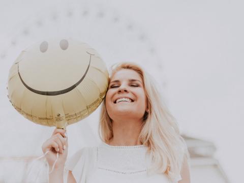 Das Bild zeigt eine glücklich scheinende Frau, die einen Luftballon mit einem Smiley in ihrer Hand hält. 