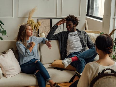 Ein Paar sitzt auf einem Sofa und diskutiert. Eine andere Frau sitzt gegenüber, hört zu und macht Notizen.